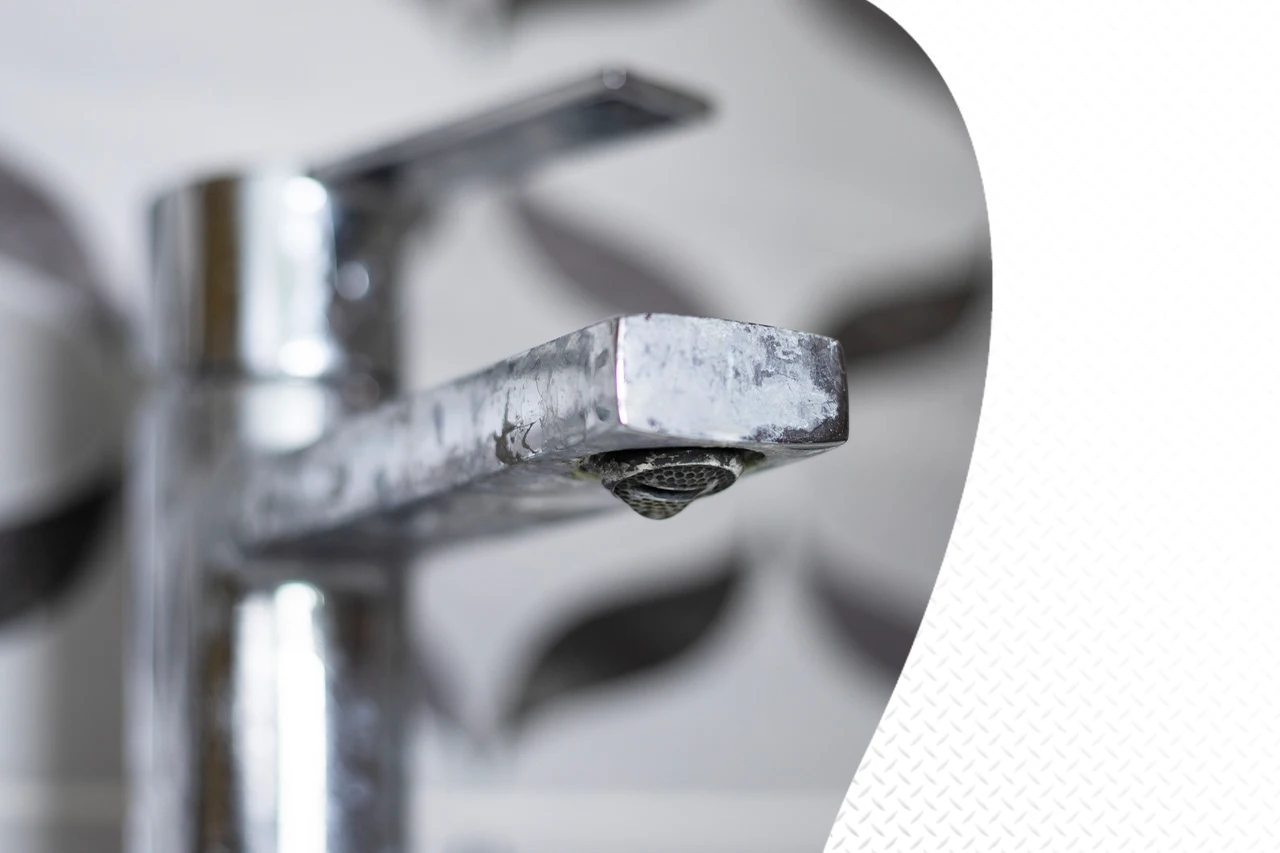 A close-up of a metallic bathroom faucet with visible limescale buildup on the spout and a single drop of water about to fall.