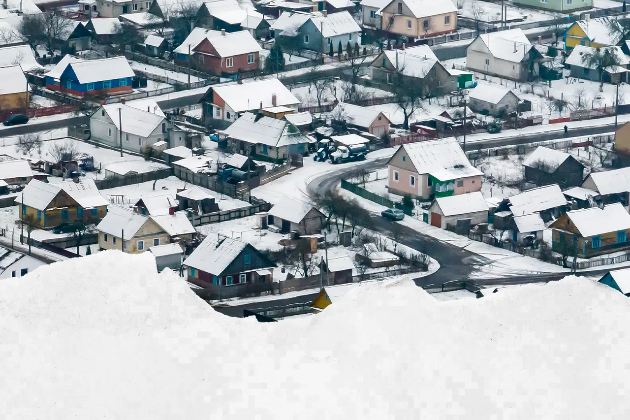 A residential neighborhood with houses and roads is covered in a light layer of snow. Snow is piled in the foreground. The scene appears calm during winter.