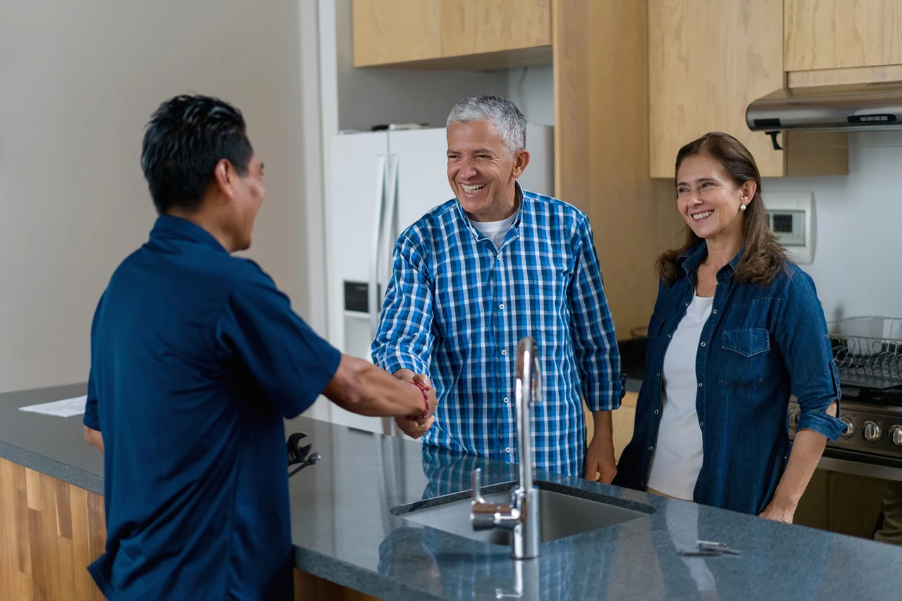 A man and woman stand in a modern kitchen while the man shakes hands with another man across the counter.