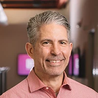 A middle-aged man with short gray hair and a trimmed beard is smiling and wearing a light red collared shirt indoors with blurred monitors in the background.