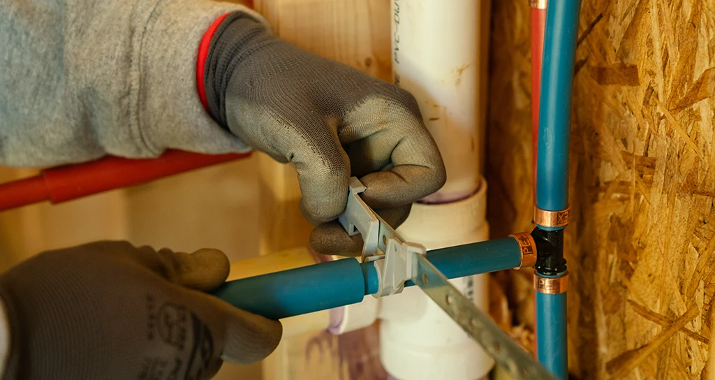 A person wearing gloves uses a pipe cutter to cut a blue plastic pipe near a plumbing system with multiple pipes and connectors.