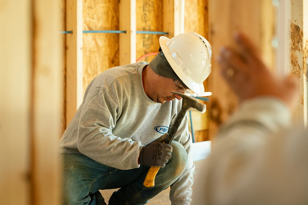 A construction worker wearing a white hard hat and gloves kneels while using a hammer inside a wooden building under construction.