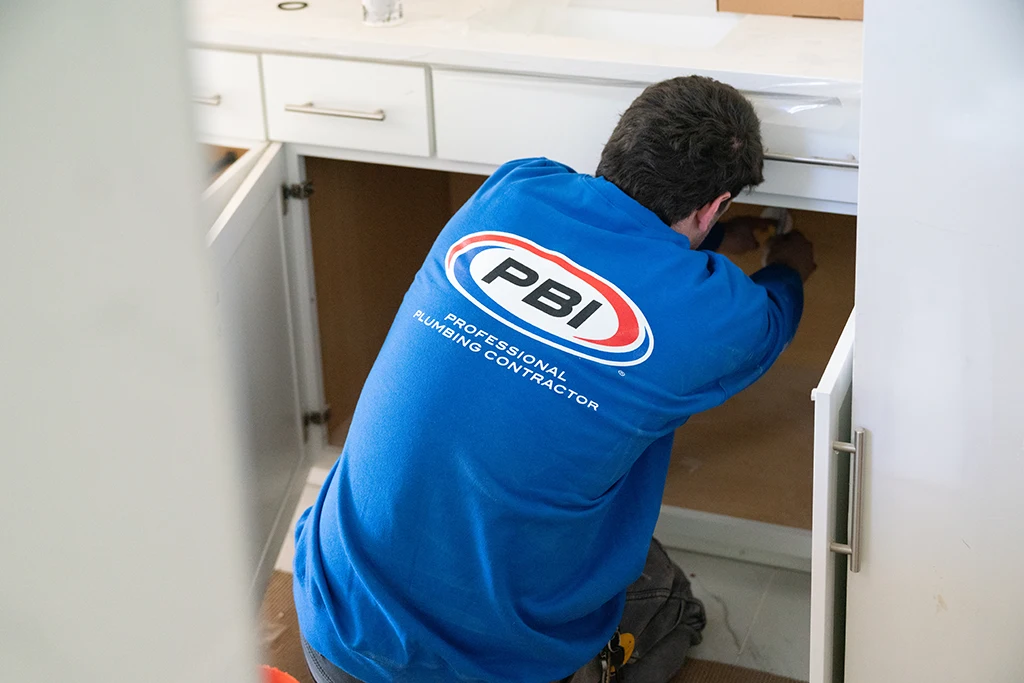 A plumber in a blue "PBI Professional Plumbing Contractor" shirt works under a bathroom sink with cabinet doors open.
