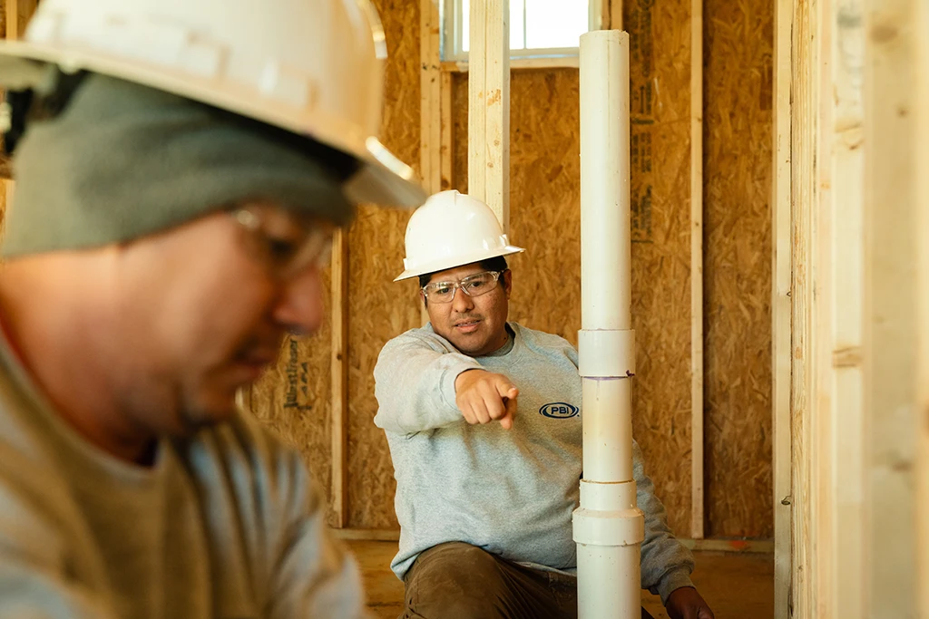 Two construction workers wearing hard hats work indoors; one is pointing towards something near a vertical PVC pipe.