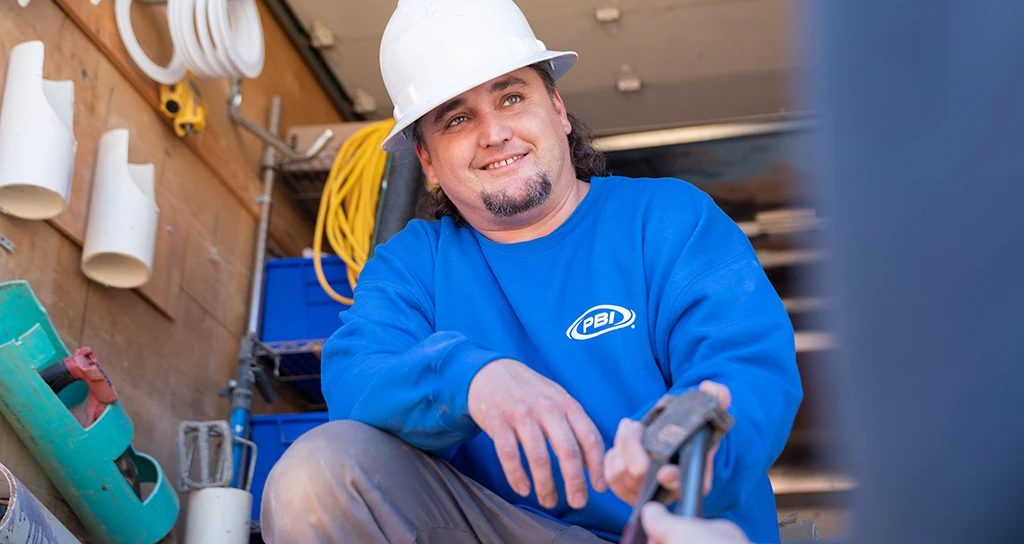 A construction worker wearing a white hard hat and blue PBI-branded sweatshirt holds a tool inside a work van with pipes and equipment visible in the background.