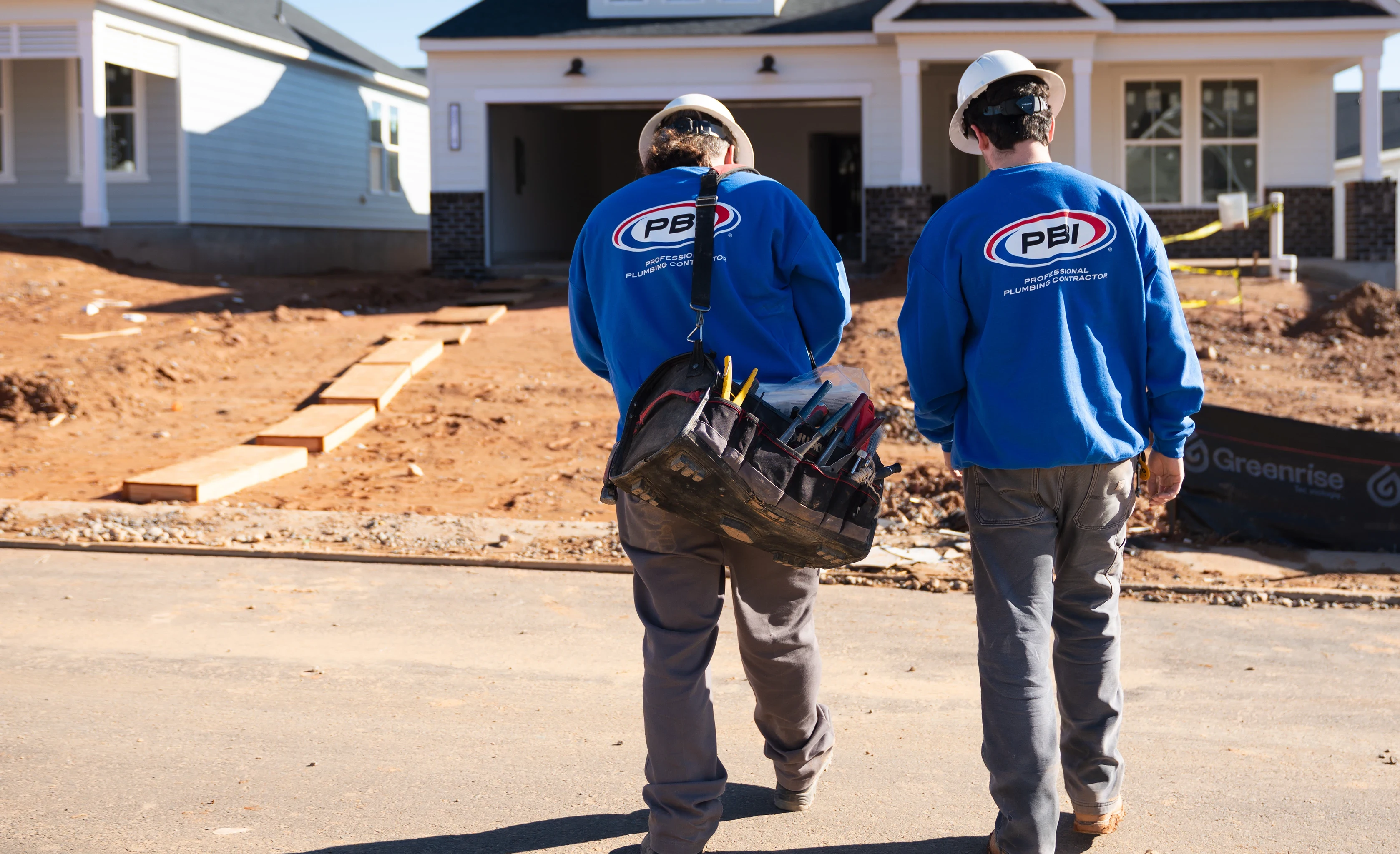 Two construction workers in blue PBI shirts and hard hats walk toward a house under construction, with one carrying a tool bag.
