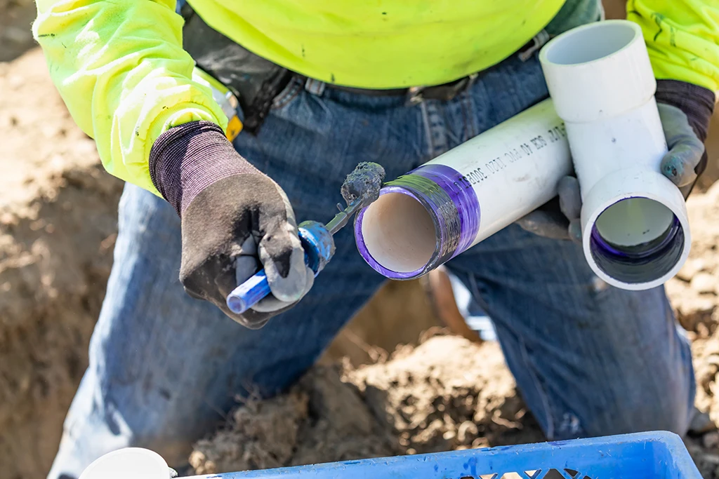 Person in work gloves applies purple PVC primer to a pipe joint with a brush, preparing to connect two white plastic pipes outdoors.