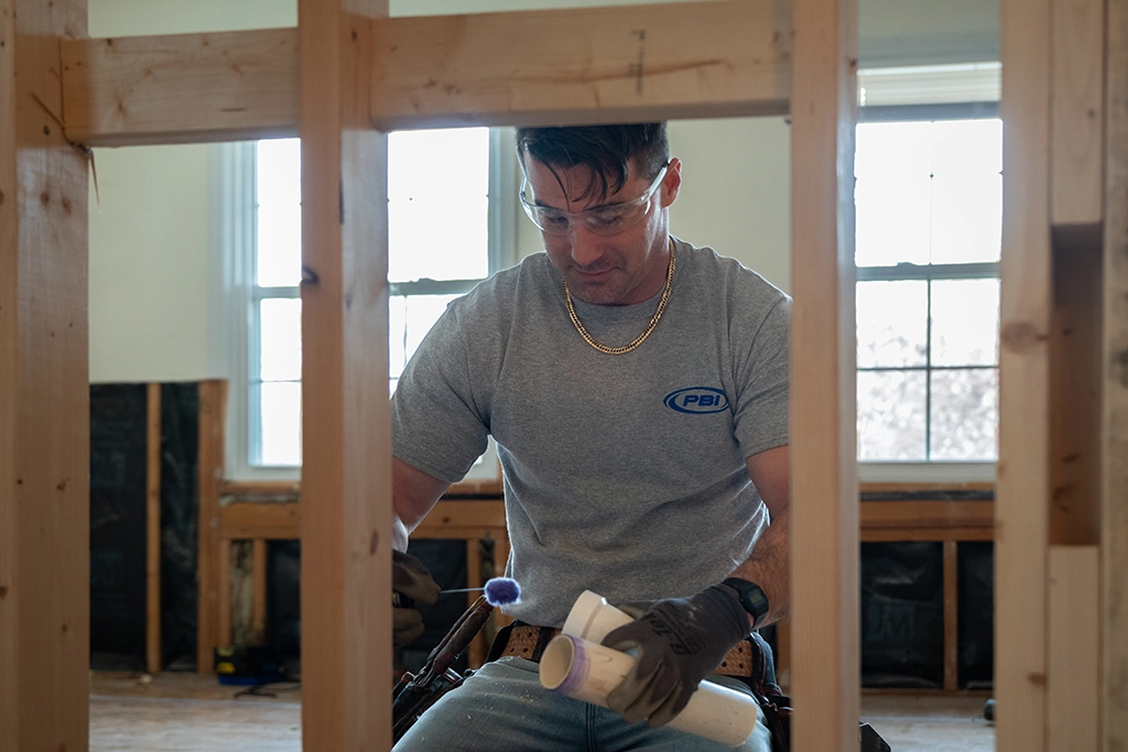 A construction worker wearing safety glasses and gloves holds PVC pipes inside a partially built wooden structure.