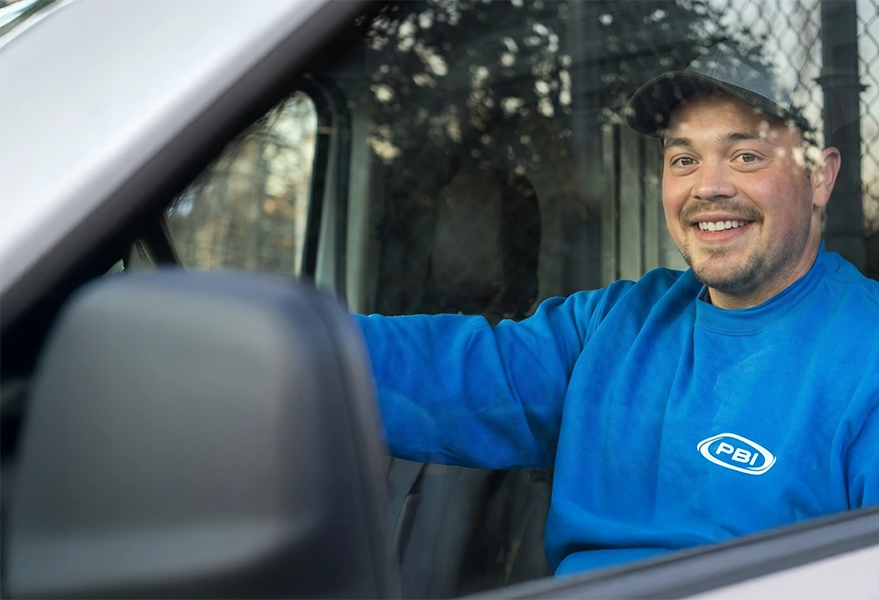 A man in a blue "PBI" sweatshirt and cap sits in the driver's seat of a vehicle, smiling at the camera through the open window.