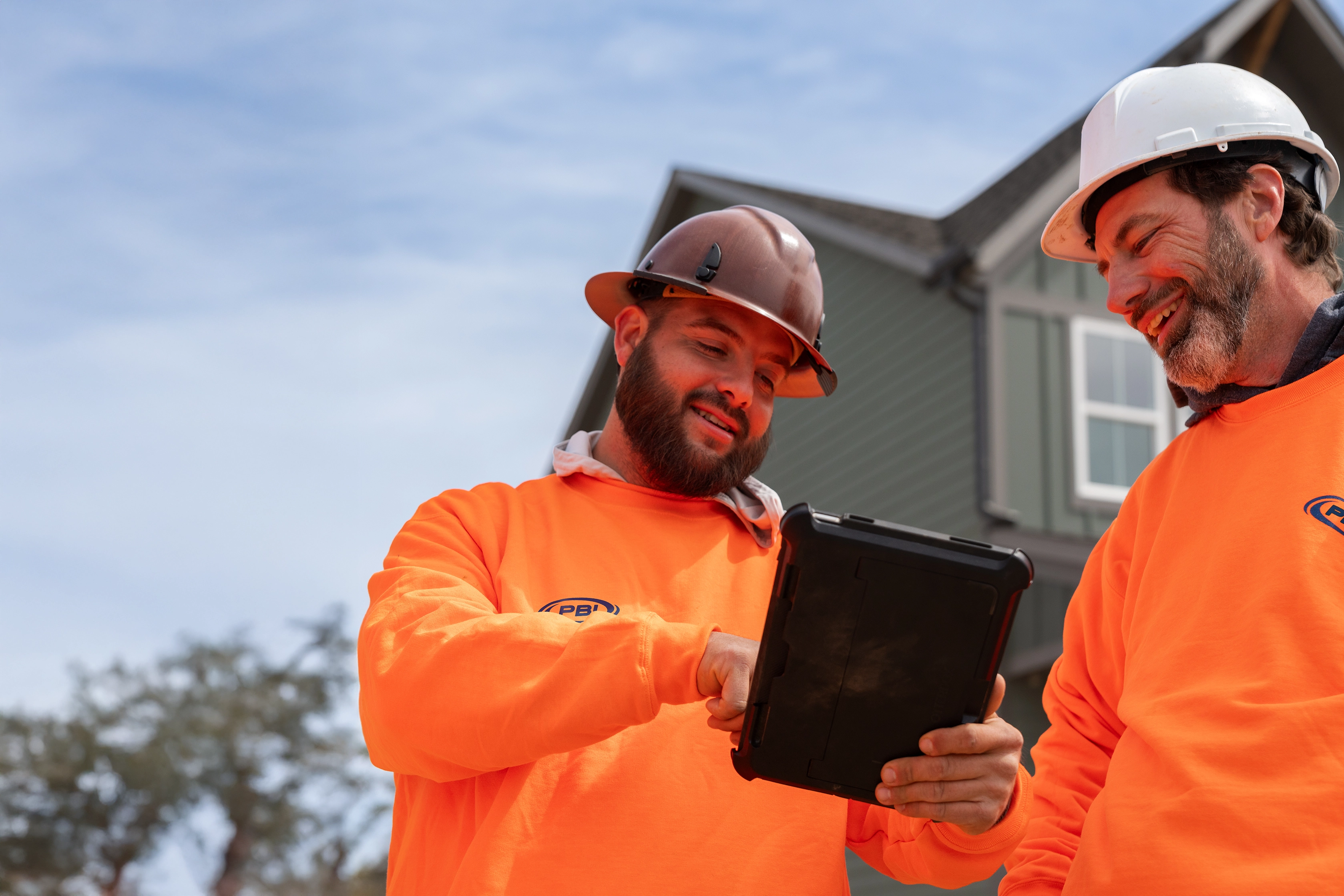 Two construction workers in orange shirts and hard hats look at a tablet outdoors in front of a house.