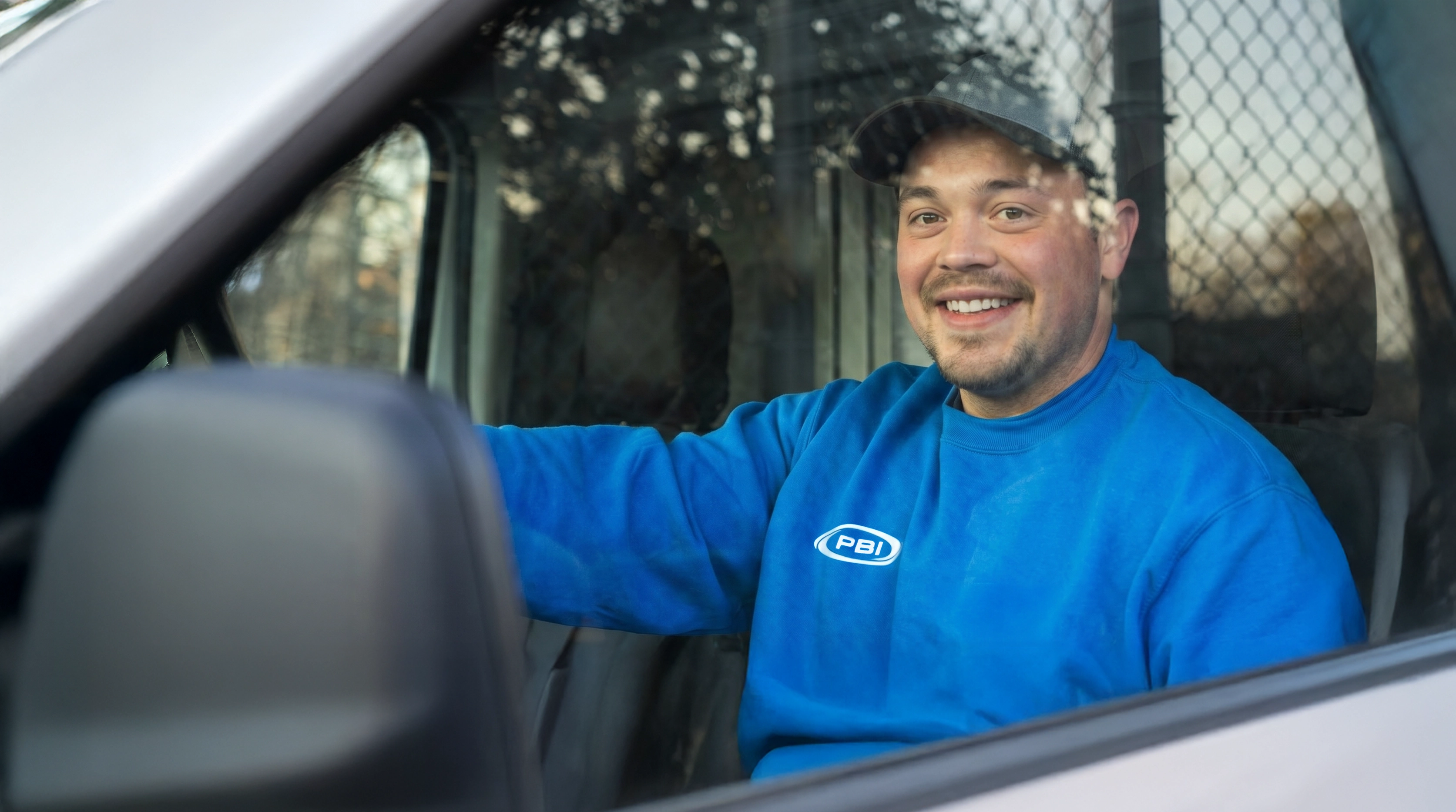 A man in a blue sweatshirt and cap smiles while sitting in the driver’s seat of a vehicle, viewed through the open window.