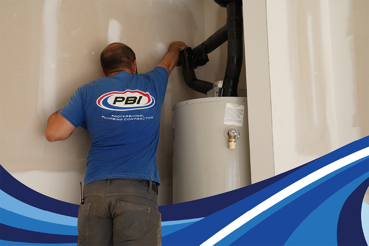 A plumber in a blue uniform working on pipes above a water heater.